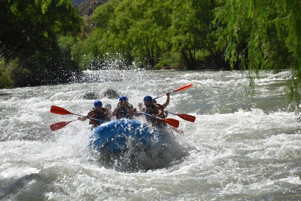 Group of rafters in a bright orange raft navigating whitewater rapids, paddles raised, surrounded by green trees.